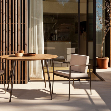 Vint Aluminium Garden Dining Armchair with beige cushions and slim tapered black legs, shown on a sunlit terrace beside a round wooden dining table.