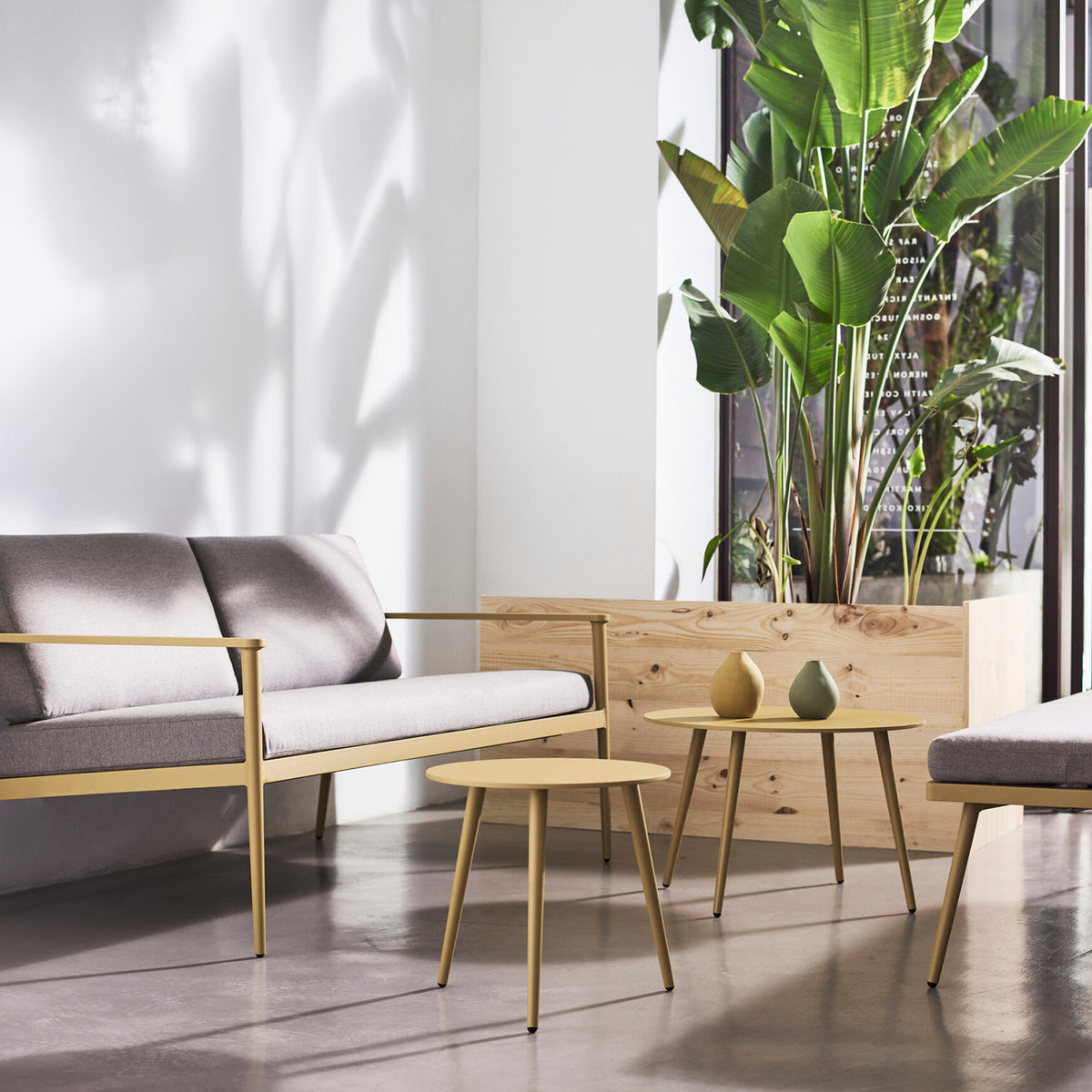 Vint Ø60cm round aluminium garden side table in mustard yellow with tapered legs, shown beside a grey sofa with two small vases on top.