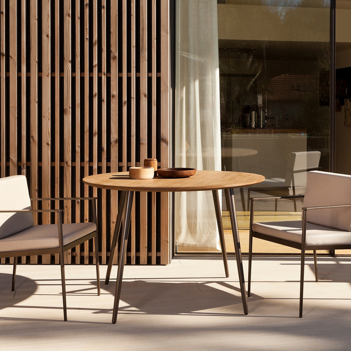 Vint Aluminium Round Outdoor Dining Table with wood-effect top and slim tapered dark metal legs, shown on a patio beside two beige outdoor armchairs.