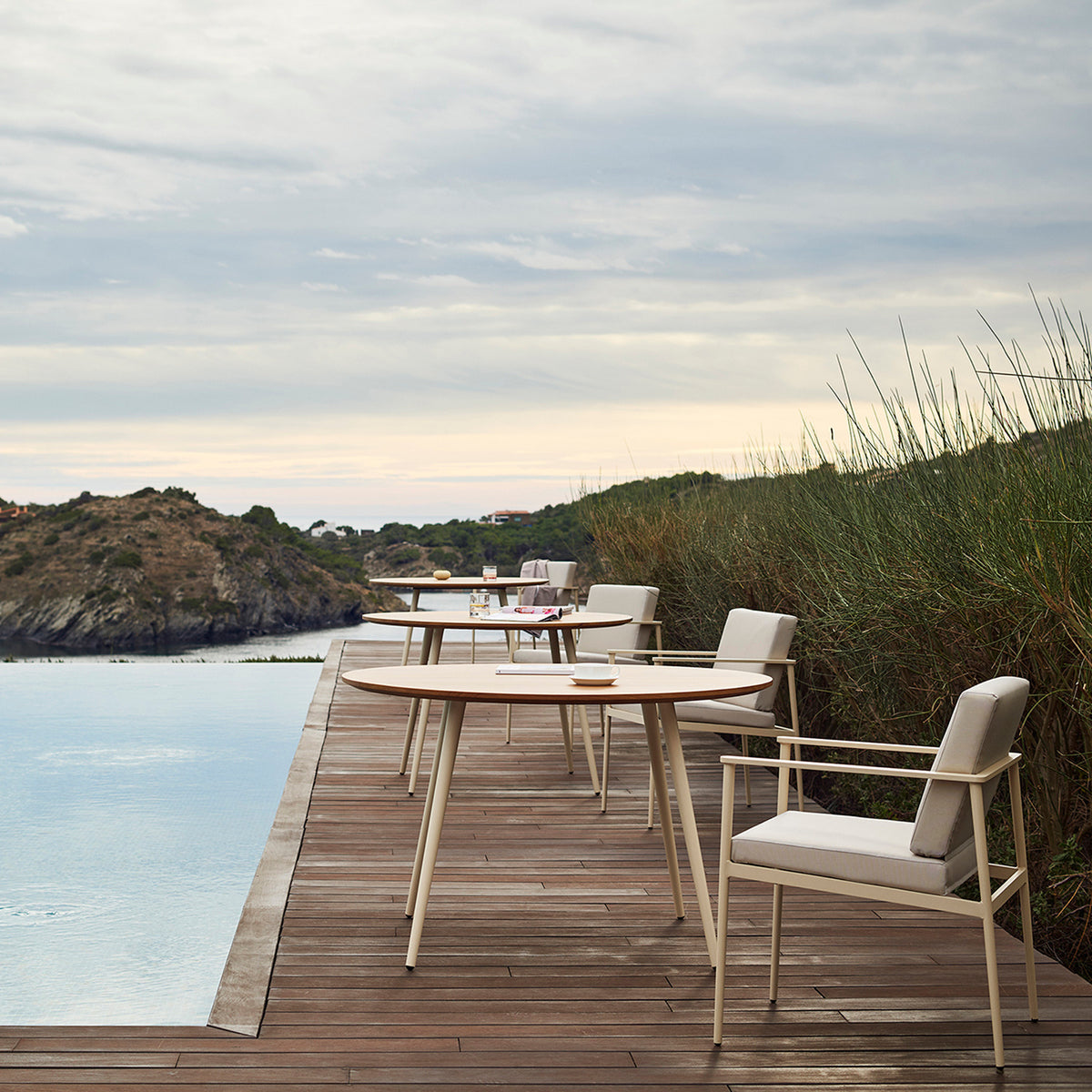 Vint Aluminium Round Outdoor Dining Tables with slim tapered legs and beige armchairs on a wooden poolside deck overlooking the sea.
