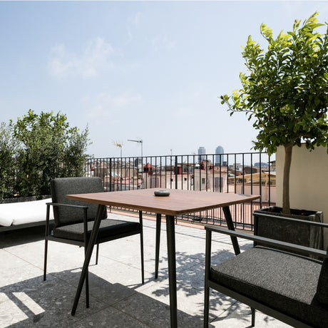 Vint Aluminium Square Outdoor Dining Table in wood-finish top with black tapered legs, shown on a rooftop terrace with armchairs, greenery and a city skyline.