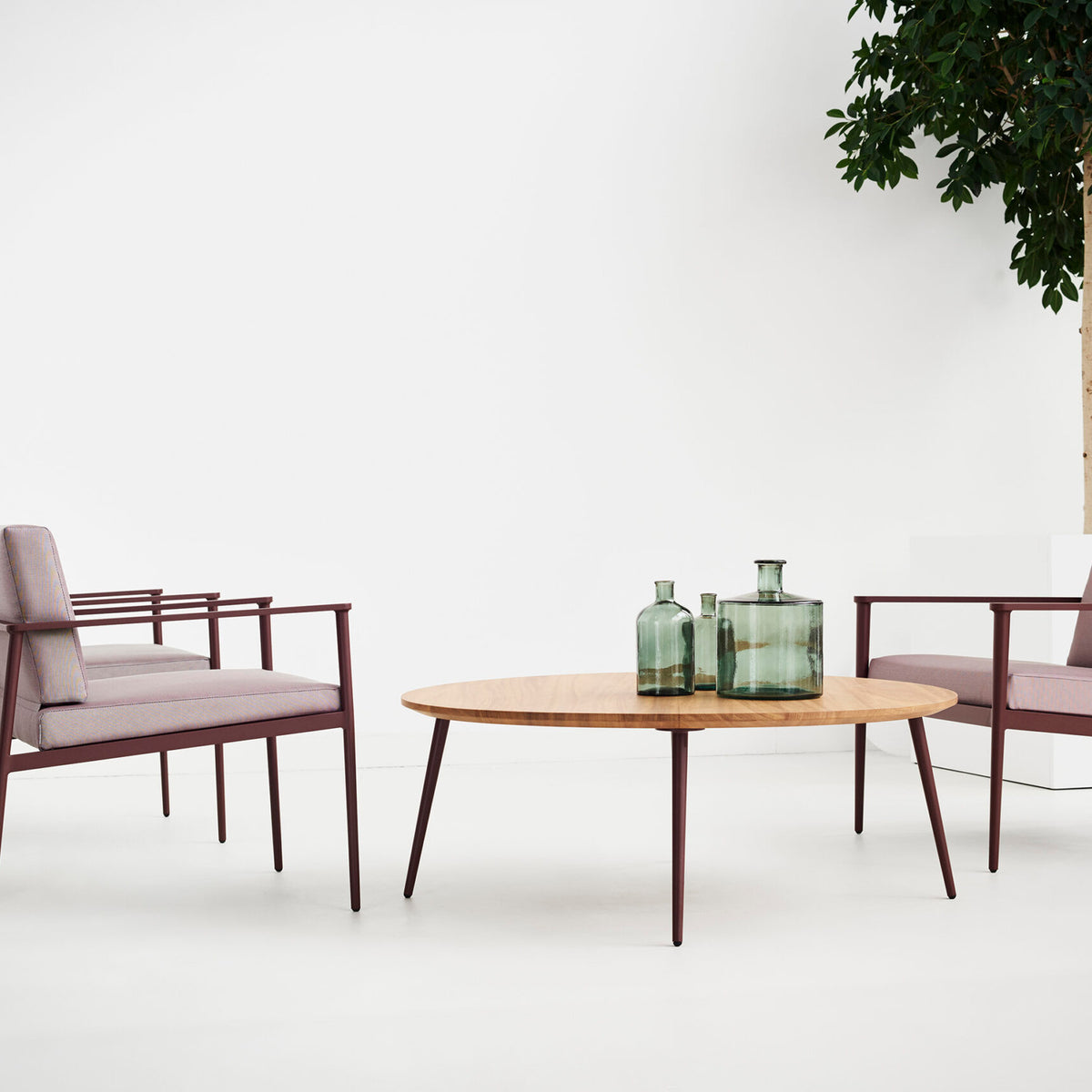 Vint round outdoor coffee table in wood-finish top with slim burgundy tapered legs, shown with two armchairs and glass vases in a light-toned room.