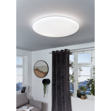 Vinvanne 76cm LED flush ceiling light with round white steel body and domed plastic diffuser, shown lit on a grey ceiling in a living room with dark curtains and a window view.