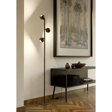 Visvette Floor Lamp in dark bronze steel with three staggered globe spotlights on a slim pole and round base, shown beside a console table in a living room.
