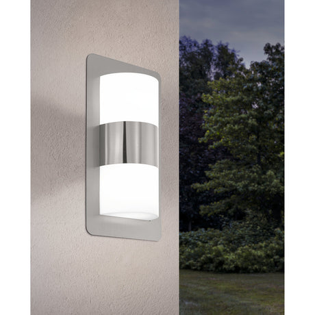 Vonvena outdoor wall light with rounded stainless steel backplate and curved white plastic shade banded by steel, lit on a textured wall beside an evening garden scene.