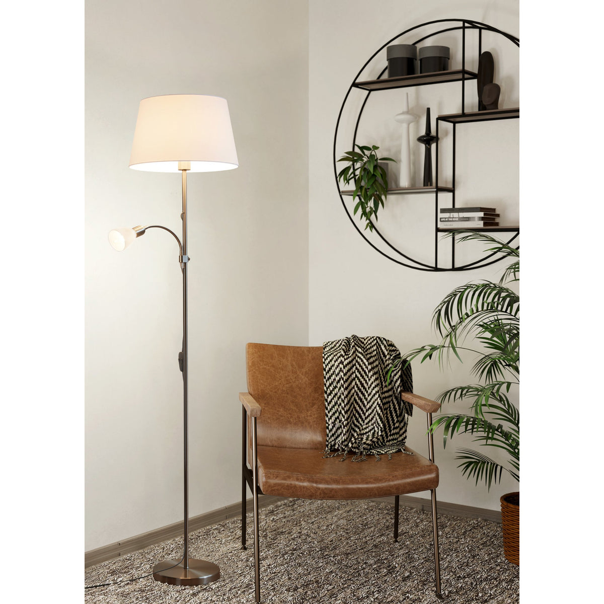 Vorbine mother and child floor lamp in satin nickel with a white tapered fabric shade and a smaller globe reading light on a gooseneck arm, shown beside a leather armchair in a living room.