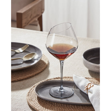 Vrede Red Wine Glass with smoke-grey tinted bowl, slanted rim, and slender stem on a round foot, filled with red wine and set on a dining table with grey plates.