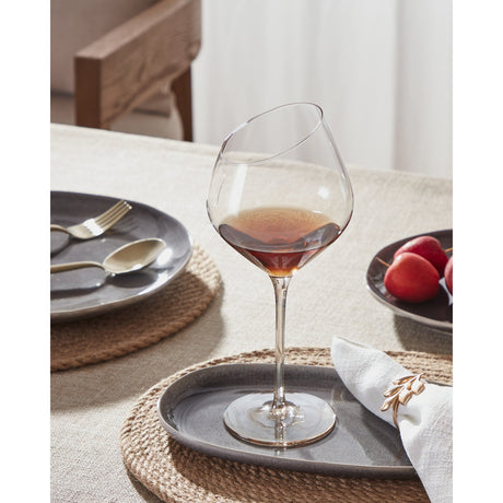 Vrede Red Wine Glass in amber-tinted glass with a slanted rim, slender stem, and round foot, shown filled on a dining table with plates and fruit.