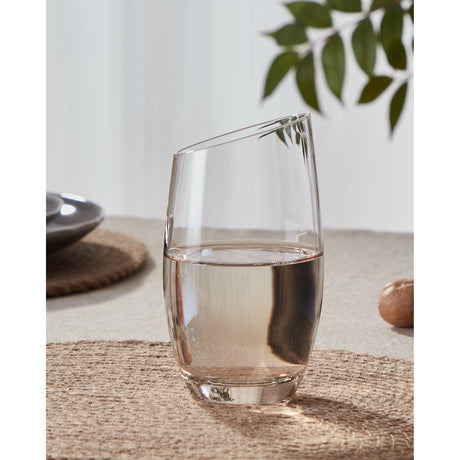 Vrede Water Glass 14.2cm in amber, a clear tumbler with a slanted rim and rounded body filled with water, set on a woven mat beside a plant and small dish.