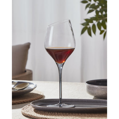 Vrede White Wine Glass with angled rim and long slender stem, holding red wine, on a grey tray at a table setting with plates and greenery in the background.