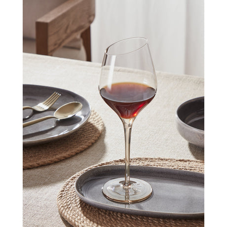 Vrede white wine glass with a tall slender stem and slanted rim, clear glass bowl warming to amber at the base, shown filled with red wine on a dining table setting.
