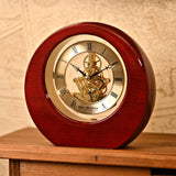 Round mantel clock in high-gloss mahogany piano wood with gold-tone dial, black Roman numerals, and an open skeleton movement showing gears, displayed on a wooden surface.