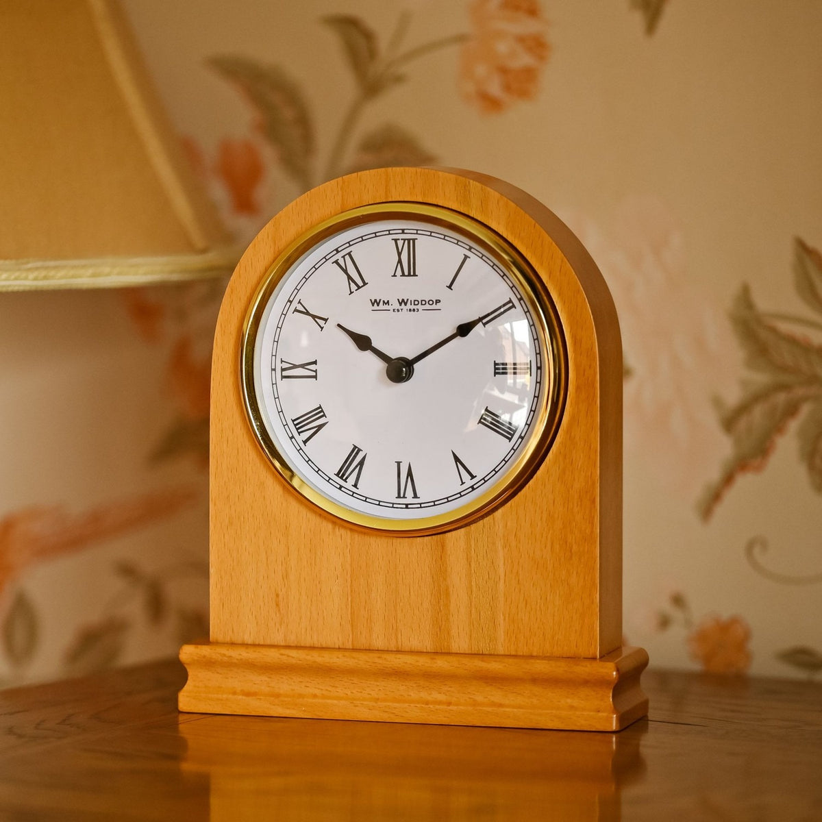 Arched oak mantel clock with rounded top, plinth base, and white Roman numeral dial in a gold-coloured bezel, shown on a dresser against floral wallpaper.