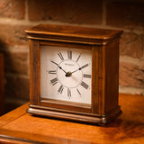 Square walnut effect Westminster chime mantel clock with white dial, black Roman numerals, spade hands, and fluted side columns, shown on a wooden surface.