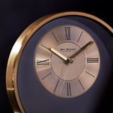 Close-up of a round gold metal mantel clock with a two-tone dial, black Roman numerals, baton markers, and slim gold hands.
