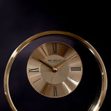 Round metal mantel clock in gold with a two-tone dial, bold black Roman numerals and baton markers, slim gold hands, set against a dark background.