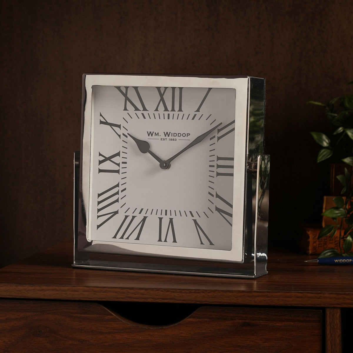 Square mantel clock with polished silver frame and white face featuring black Roman numerals, displayed on a wooden sideboard in a living room setting.