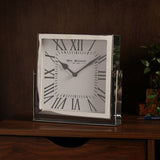 Square mantel clock with polished silver frame and white face featuring black Roman numerals, displayed on a wooden sideboard in a living room setting.