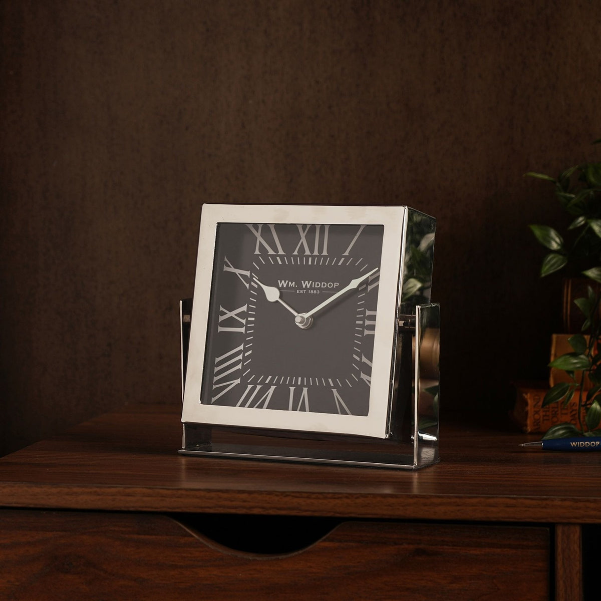 Square mantel clock in silver frame with black dial, white Roman numerals, held on a matching silver stand on a dark wood sideboard.