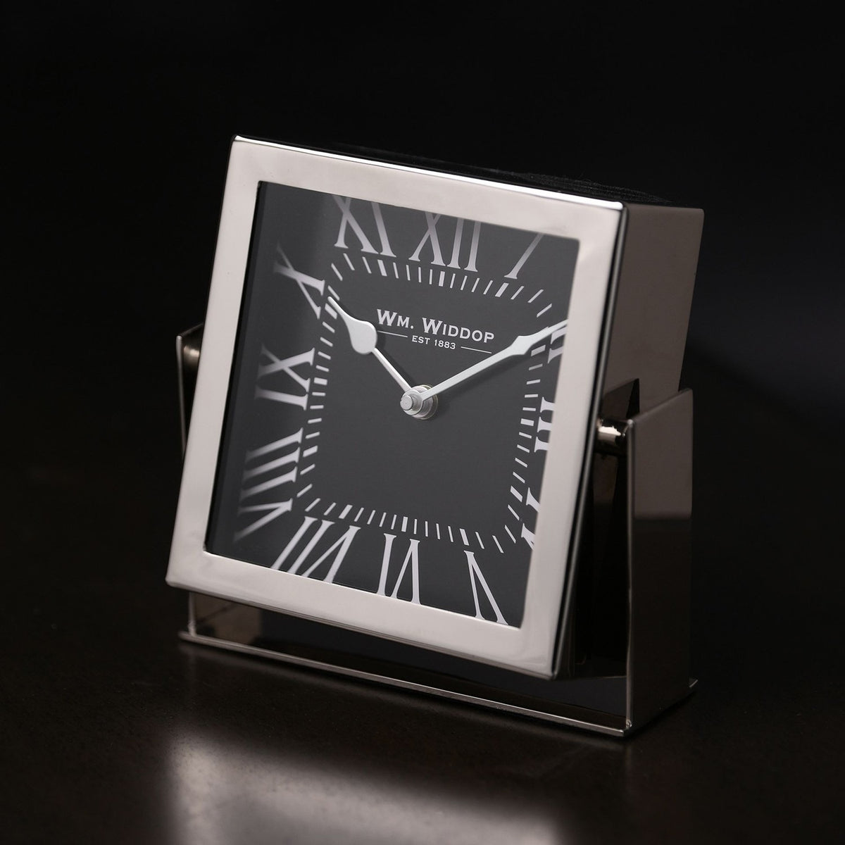 Square mantel clock with silver frame and stand, black dial, white Roman numerals and hands, shown at an angle against a dark background.