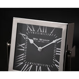 Close-up of a square mantel clock with silver frame and stand, black dial, white Roman numerals, and silver hands, set against a dark background.