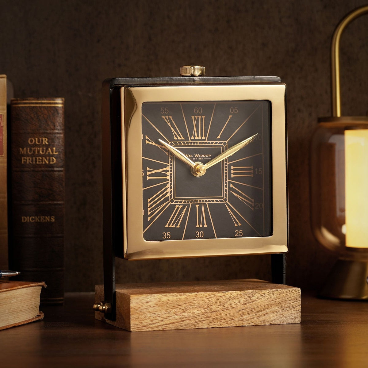 Square mantel clock with black Roman numeral dial in a polished gold case, set between black uprights on a wooden base, shown beside books on a desk.
