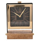 Square mantel clock with black Roman numeral dial in a polished gold case, set between black uprights on a wooden rectangular stand, shown on a plain white background.