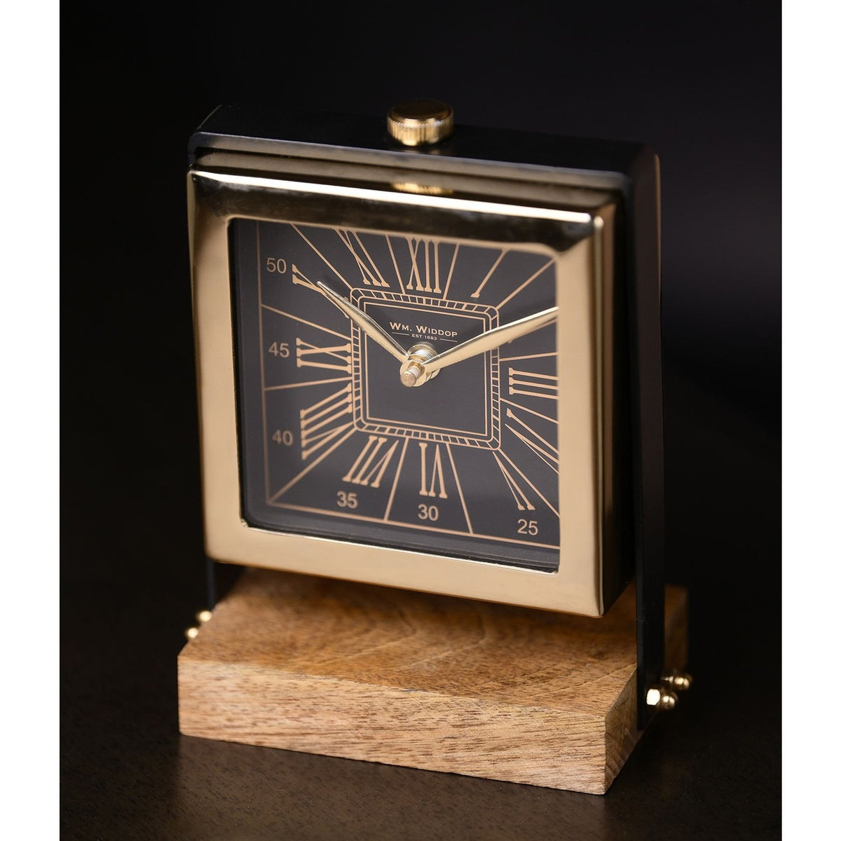 Square mantel clock with black Roman numeral dial in a polished gold case, set between black uprights on a wooden table stand, against a dark background.