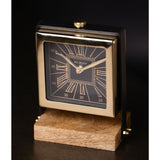 Square mantel clock with black Roman numeral dial in a polished gold case, set between black uprights on a wooden table stand, against a dark background.