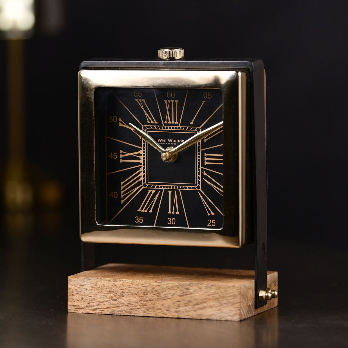 Square mantel clock with black dial and gold Roman numerals in a polished gold case, mounted on black uprights above a wooden rectangular base, dark background.