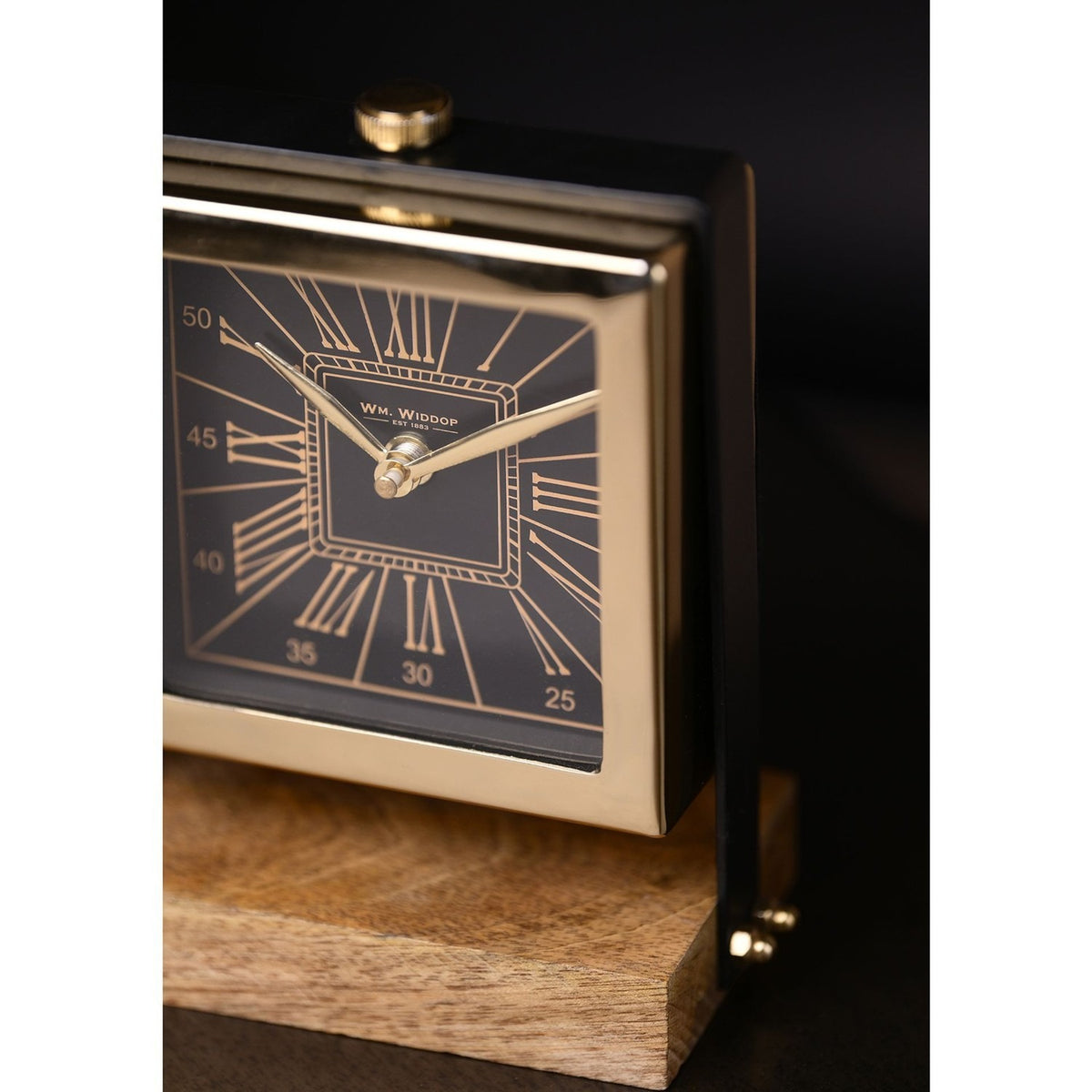 Close-up of the Square Roman Numeral Mantel Clock with a black dial, gold Roman numerals, minute track, and gold hands set in a polished gold case on a wooden stand.
