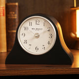 Napoleon resin mantel clock in matt black with arched hat-shaped silhouette, round white dial, silver rim, black Arabic numerals, and slim silver hands, shown on a wooden surface beside books.