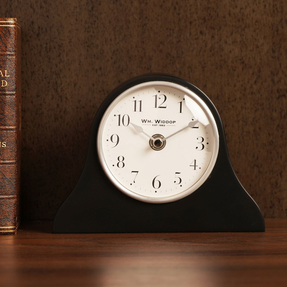 Napoleon arch mantel clock in black resin with matt finish, round white dial, silver rim, black Arabic numerals and slim silver hands, shown beside a leather-bound book.