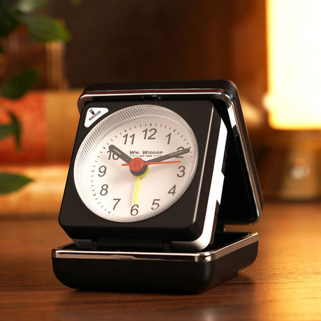 Mini square alarm clock in a black folding case with silver trim, opened to show a white dial with bold numerals, black hands, red seconds hand and yellow alarm hand, displayed on a wooden surface.