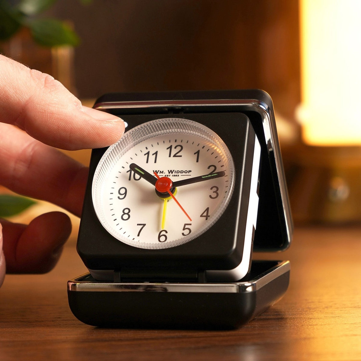 Mini square alarm clock in black folding case, opened to show white dial with bold numerals, black hands, red seconds hand and yellow alarm hand, held by a hand on a wooden bedside table.