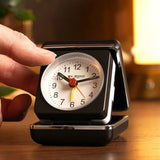 Mini square alarm clock in black folding case, opened to show white dial with bold numerals, black hands, red seconds hand and yellow alarm hand, held by a hand on a wooden bedside table.