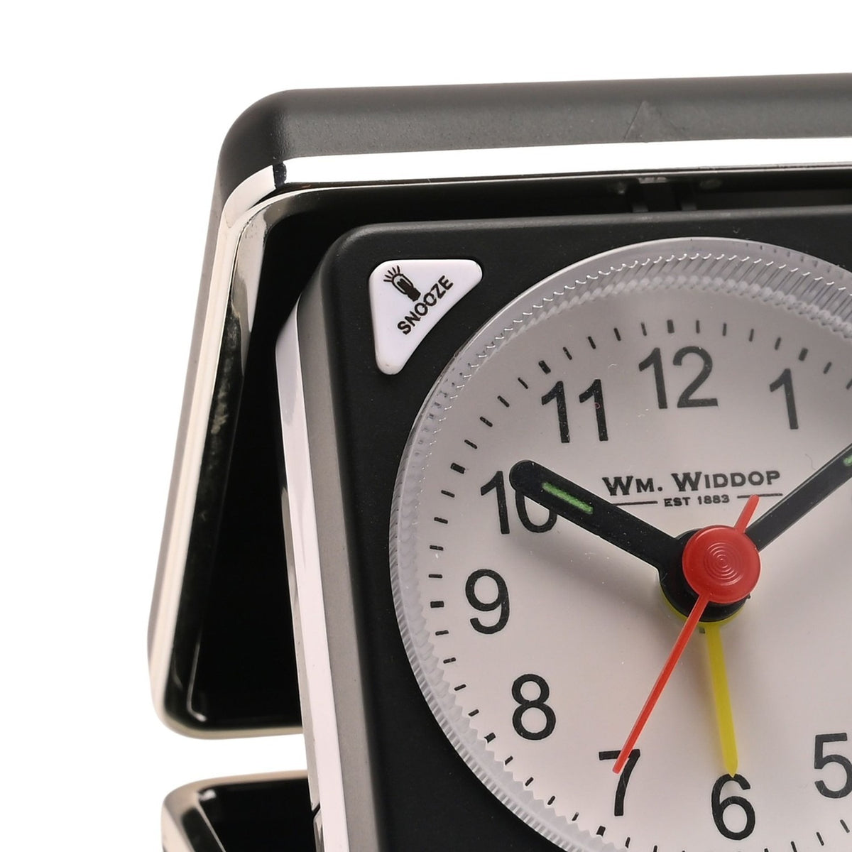 Close-up of the mini square alarm clock in black folding case with silver trim, showing white dial, black numerals, snooze button, and red and yellow hands.