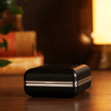 Mini square alarm clock in its black folding case with a silver edge trim, shown closed on a wooden surface in a softly lit room setting.
