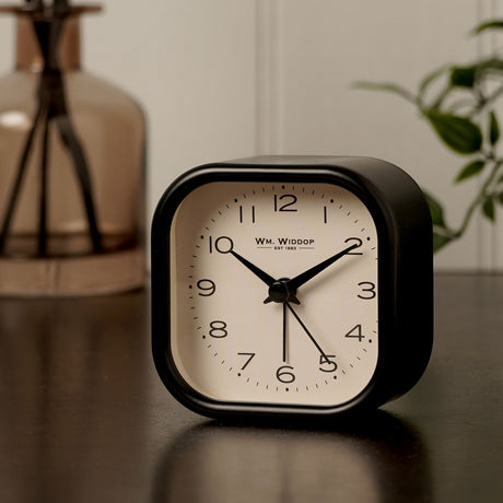 Rounded square alarm clock with black metal case and cream dial, bold Arabic numerals, and slender black hands, shown on a dark surface beside a plant and glass vase.