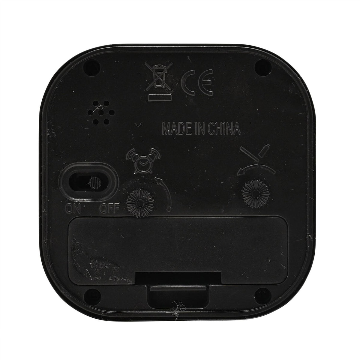 Rear view of the rounded square black metal alarm clock showing on/off switch, alarm set and time adjustment wheels, speaker holes, and battery compartment on a white background.