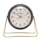 Medium Kent round metal alarm clock in matt black with a white dial, gold hands, and numbers one to twelve, set on a slim gold wire stand against a plain white background.