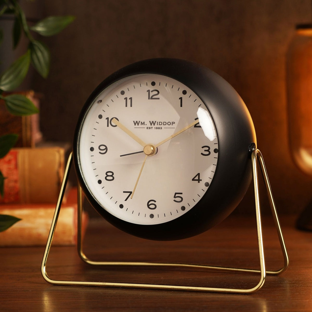 Medium Kent round metal alarm clock in matt black with a white dial, gold hands, and a slim gold wire stand, shown on a wooden surface.