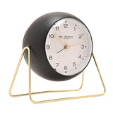 Round black metal alarm clock with white dial, gold hands, and clear numbers, set on a slim gold wire stand against a plain white background.