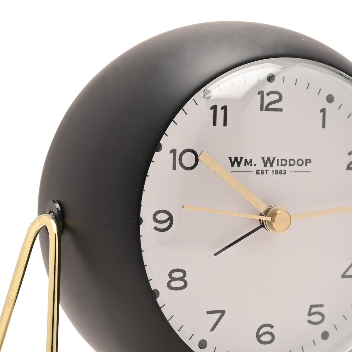 Close-up of the Medium Kent round metal alarm clock in matt black with a white dial, clear numbers, dot hour markers, gold hands and a domed cover, set on a slim gold wire stand.
