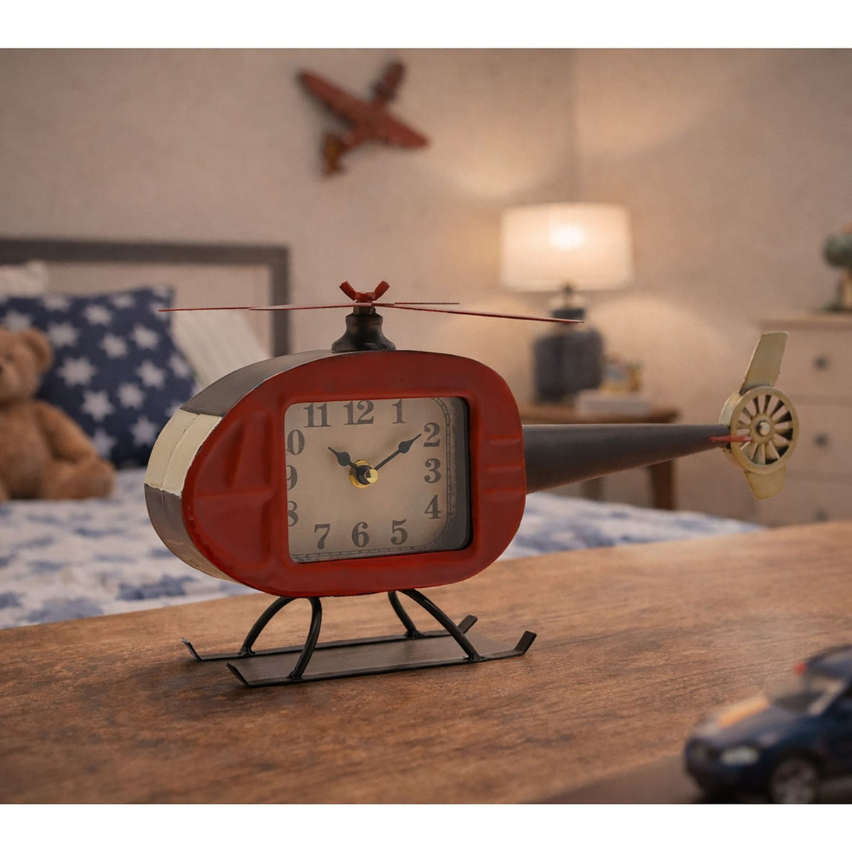 Helicopter Metal Mantel Clock with red rounded cabin, cream dial with black numerals, black tail boom, rotor blades and landing skids, displayed on a table in a bedroom setting.