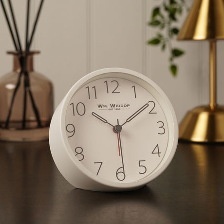 Round white metal analogue alarm clock with bold black numerals, grey hour and minute hands and a red second hand, displayed on a dark tabletop with a plant and lamp in the background.