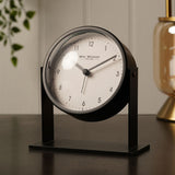 Round Adjustable Metal Alarm Clock, 13.5cm, with white dial and matt black frame on an upright stand, shown on a desk beside a plant and lamp.