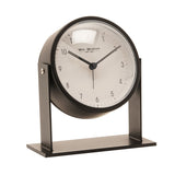 Round adjustable alarm clock with white dial and black numerals, set in a matt black metal case tilted between two upright arms on a rectangular base, shown on a plain white background.
