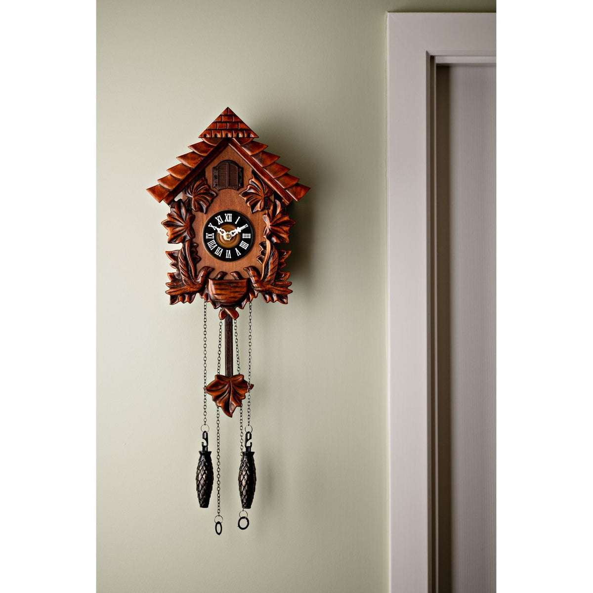 Mahogany cuckoo wall clock in a pitched-roof house shape with carved leaves, a shuttered door, a black Roman numeral dial, and hanging chain-weight pendulums, mounted beside a doorframe.
