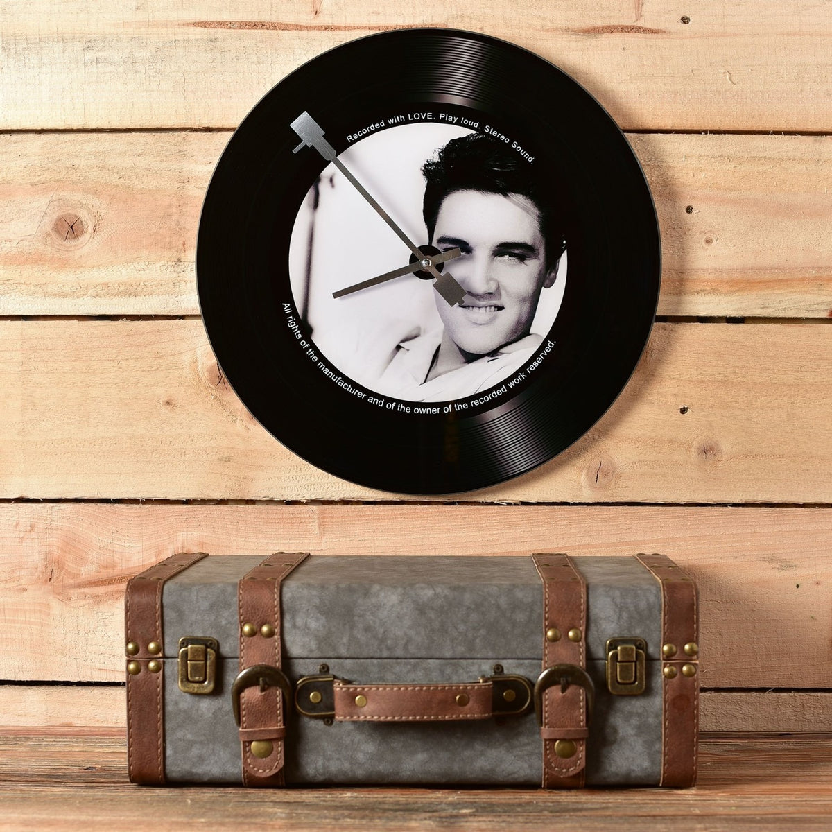Round black glass wall clock styled as a vinyl record, 37.9cm, with grooved rim, portrait centre label, and needle hands, mounted above a suitcase against a wood panel wall.